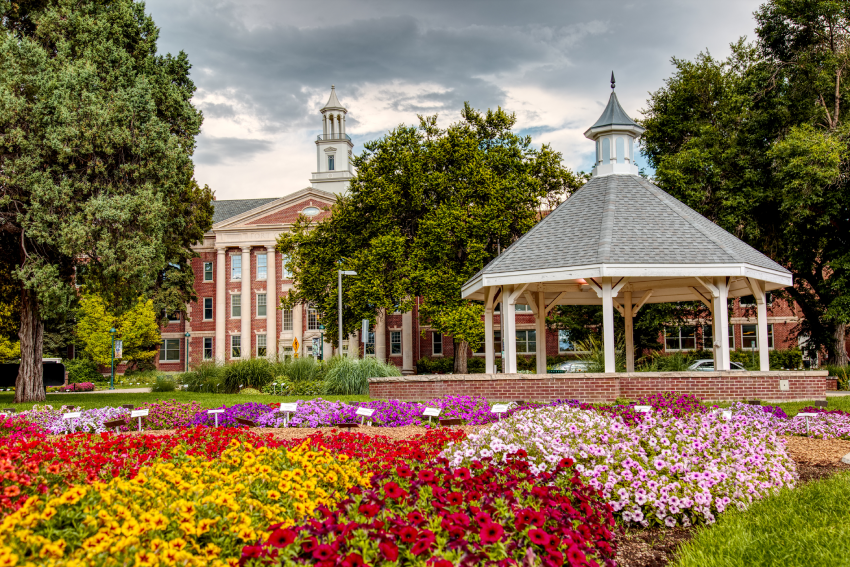 A gazebo and a bed of colorful flowers in front of a brick building with a tower and clock, surrounded by trees under a cloudy sky. A gazebo and a bed of colorful flowers in front of a brick building with a tower and clock, surrounded by trees under a cloudy sky.