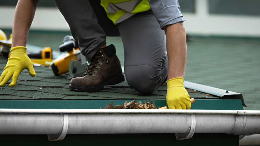 Person in gloves cleaning leaves from a gutter on a roof, with tools nearby. Person in gloves cleaning leaves from a gutter on a roof, with tools nearby.