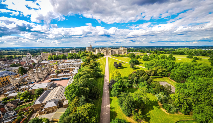 Aerial view of a castle surrounded by expansive lawns, trees, and nearby buildings under a partly cloudy sky. Aerial view of a castle surrounded by expansive lawns, trees, and nearby buildings under a partly cloudy sky.