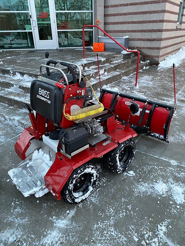 A red snow removal machine with the label “Boss” is parked on a snow-covered concrete surface near a building entrance and stairs. An orange shovel leans against the railing nearby. A red snow removal machine with the label "Boss" is parked on a snow-covered concrete surface near a building entrance and stairs. An orange shovel leans against the railing nearby.