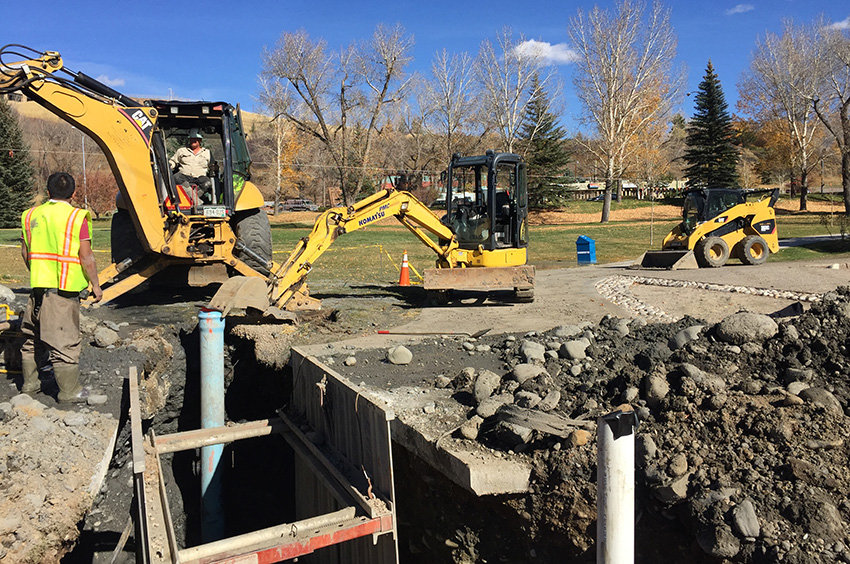 Construction workers operate excavators and machinery near a dug trench at a site with dirt, rocks, and pipes, surrounded by leafless trees. Construction workers operate excavators and machinery near a dug trench at a site with dirt, rocks, and pipes, surrounded by leafless trees.