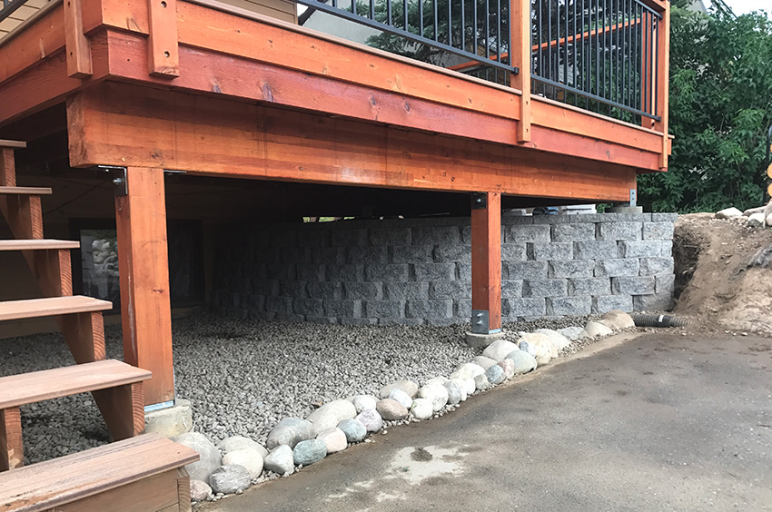 Wooden deck supported by beams with a stone retaining wall underneath. Gravel and rocks are on the ground, and a concrete surface is in the foreground. Wooden deck supported by beams with a stone retaining wall underneath. Gravel and rocks are on the ground, and a concrete surface is in the foreground.