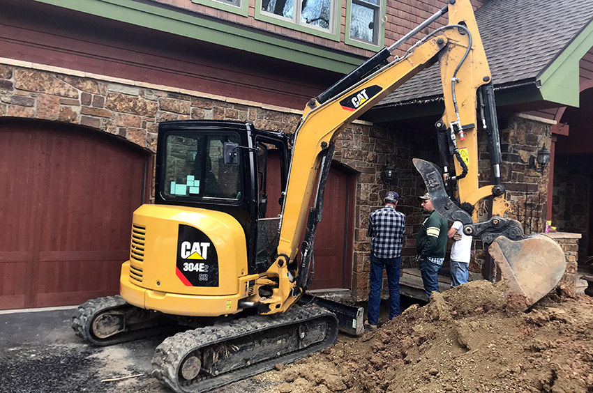 Construction workers use a yellow excavator next to a brick building with large wooden doors.