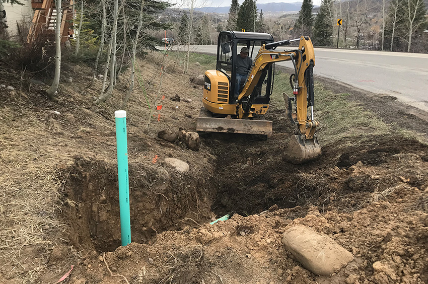 A small excavator digs a trench by the roadside, with a blue pipe visible in the ground and trees in the background.