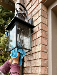 A person uses a blue cloth to clean a glass outdoor lantern mounted on a brick wall. A person uses a blue cloth to clean a glass outdoor lantern mounted on a brick wall.