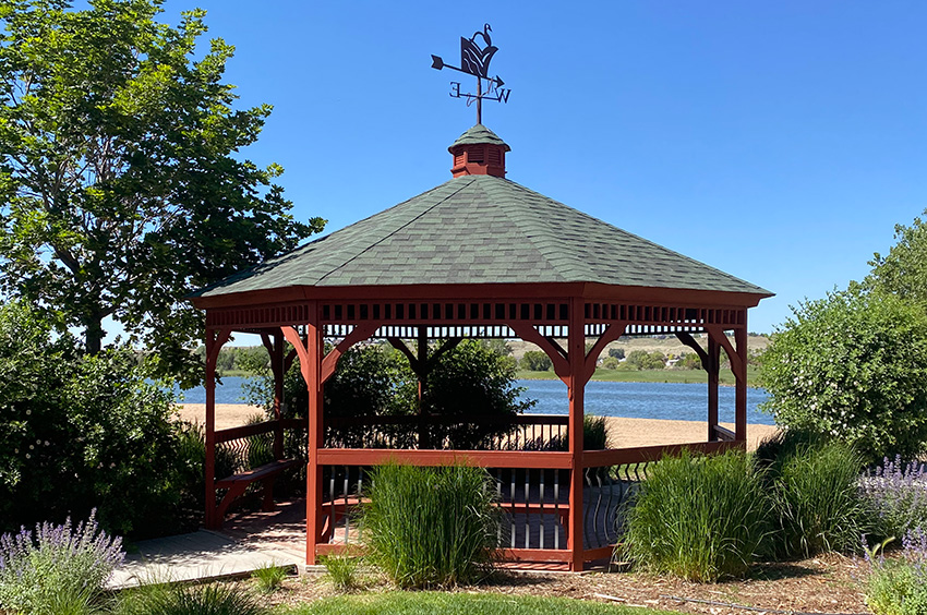 A wooden gazebo with a green roof and weather vane stands near a body of water, surrounded by greenery and a clear blue sky.