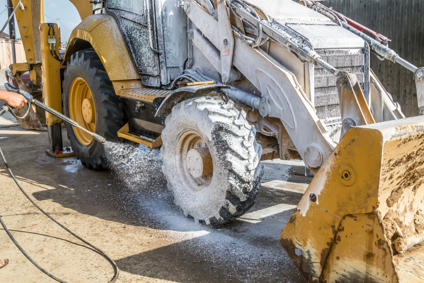 A worker uses a pressure washer to clean the muddy wheel of a yellow backhoe loader at a construction site.