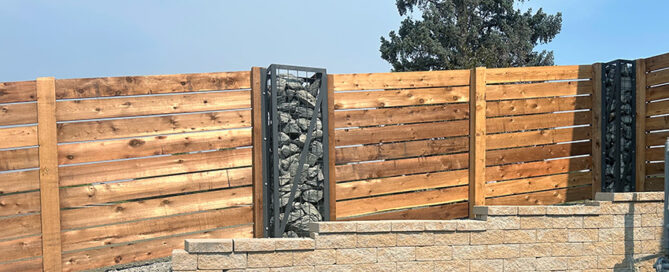 A wooden fence with stone-filled metal posts is set atop a short stone wall. A tree and gravel ground are visible in the background.