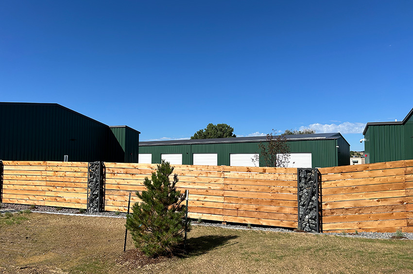 A wooden fence with stone pillars stands in front of green metal buildings under a clear blue sky. A small pine tree is in the foreground.