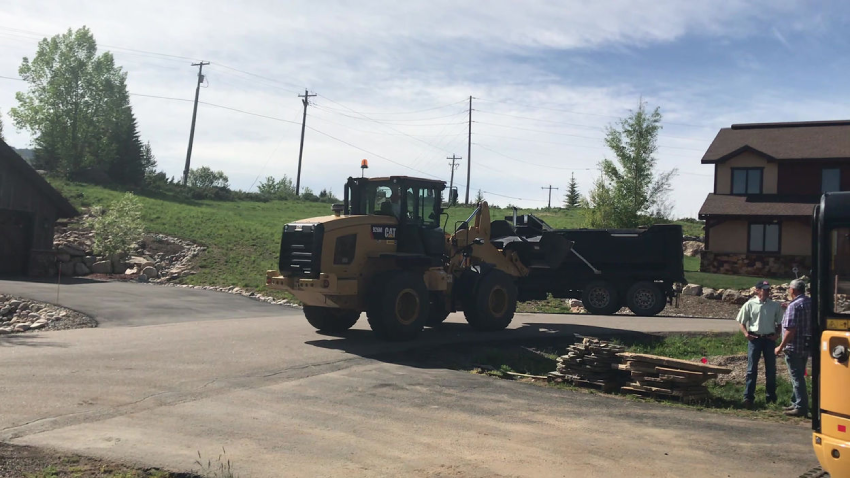 Construction vehicle maneuvering near a house and dump truck on a paved driveway with two people nearby.