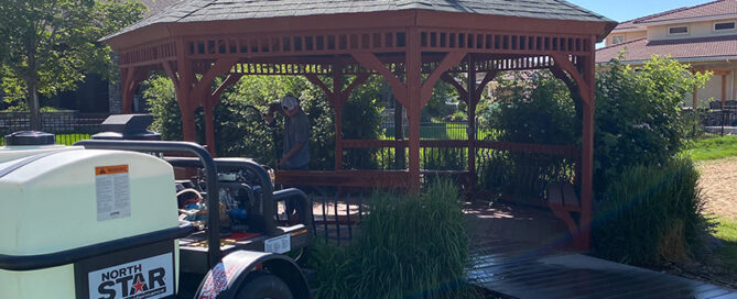 A person stands inside a wooden gazebo surrounded by greenery. A trailer with a white tank labeled "North Star" is parked nearby. Houses and trees are in the background.