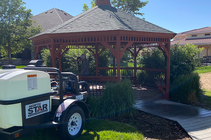 A person stands inside a wooden gazebo surrounded by greenery. A trailer with a white tank labeled "North Star" is parked nearby. Houses and trees are in the background.