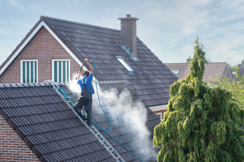 Worker in blue overalls using a high-pressure hose to clean a tiled roof on a two-story brick house, standing on a ladder with steam rising, near a large tree.