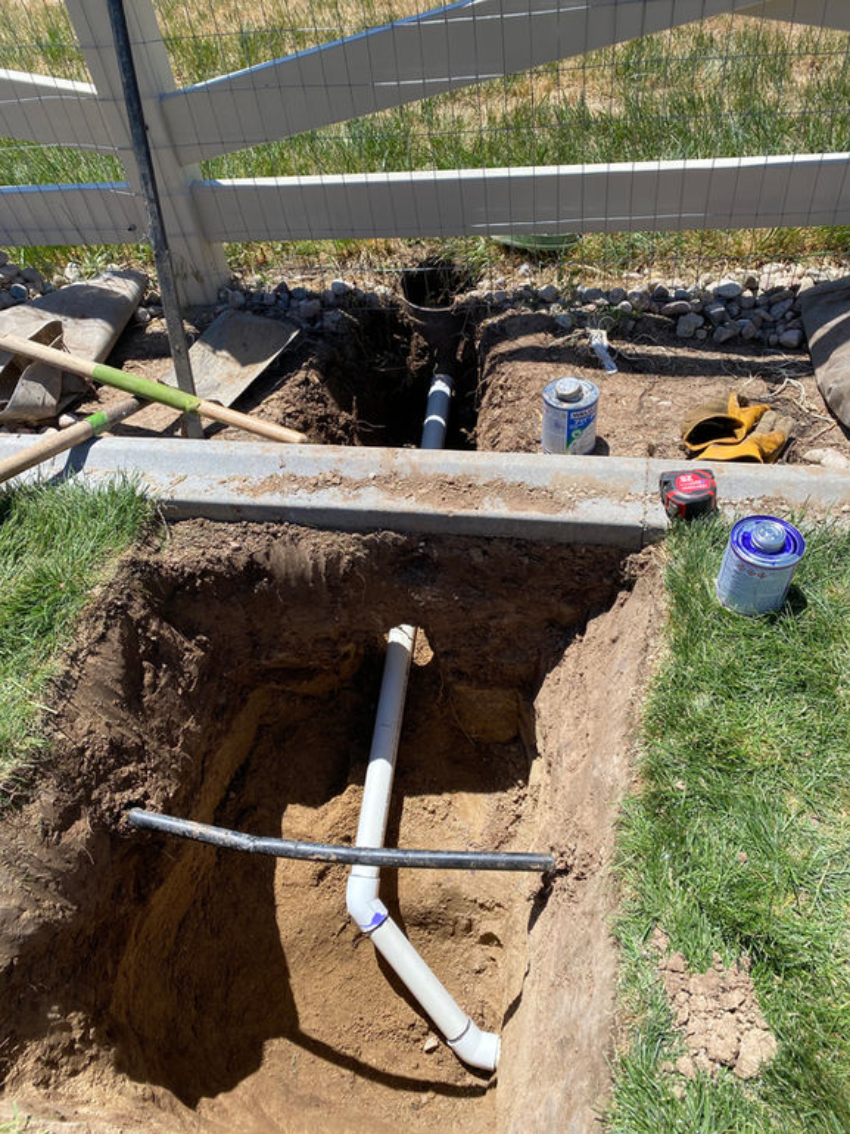 Trench with PVC pipes laid underground near a white fence. Soil piles and tools are visible alongside paint cans.