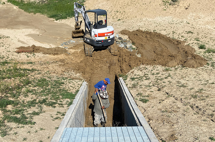 Two workers in a trench; one operates a mini excavator, while the other stands nearby. The scene is on a dirt construction site with a metal grate bridge overhead.