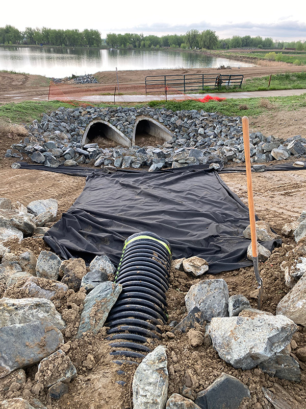 Construction site showing a drainage system with two culverts and black sheeting, surrounded by rocks. A shovel is positioned nearby, and a pond is visible in the background. Construction site showing a drainage system with two culverts and black sheeting, surrounded by rocks. A shovel is positioned nearby, and a pond is visible in the background.