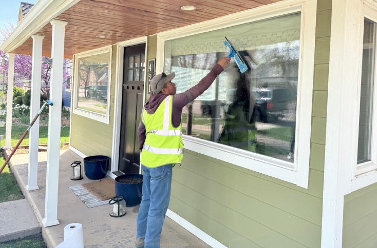 A person wearing a safety vest cleans the large front window of a green house using a squeegee. Cleaning supplies are placed on the porch nearby.